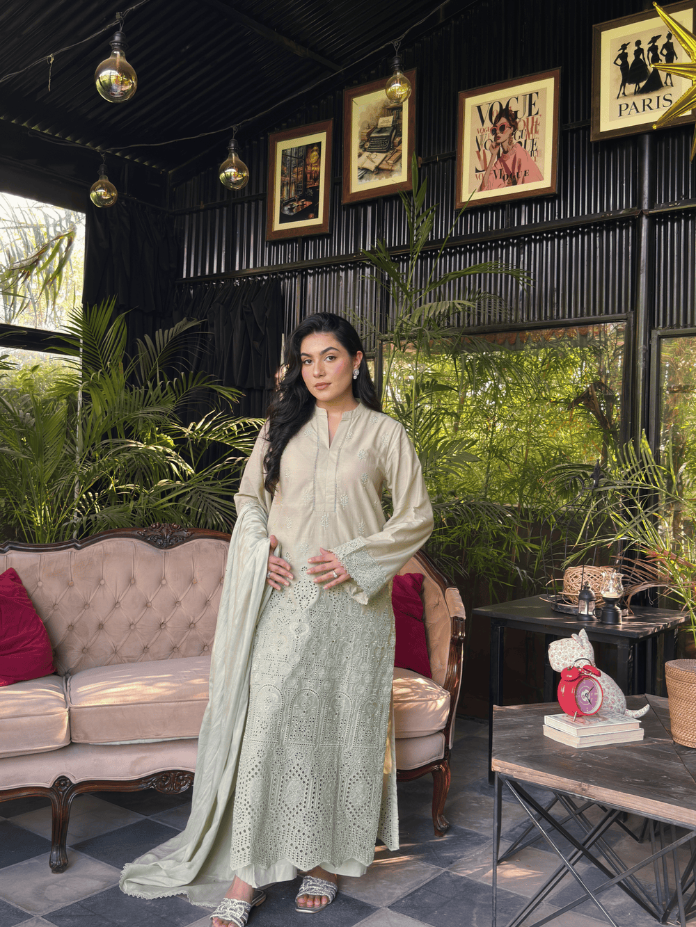 A woman models a light sage green traditional outfit with matching dupatta in a styled indoor setting with a pink sofa, potted plants, and framed wall art.
